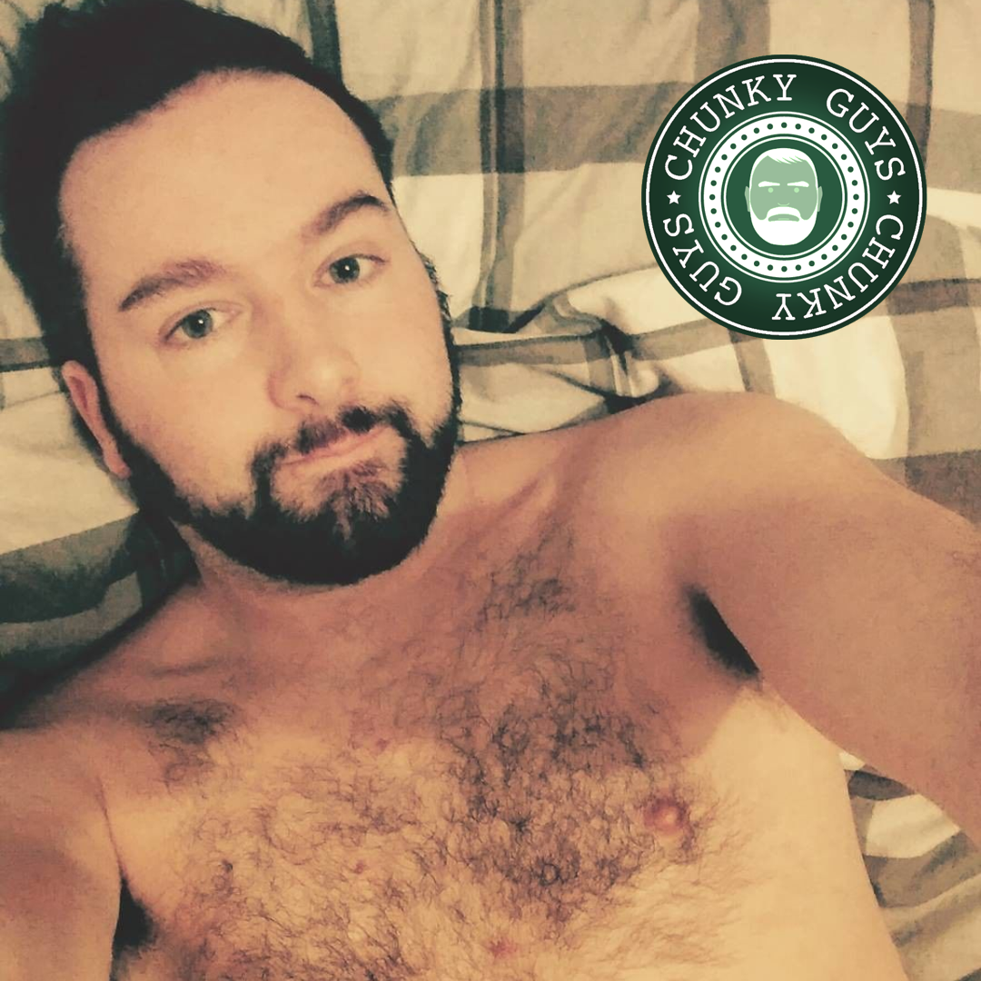 Beadrded guy with hairy chest lying down on a bed, looking up at the camera