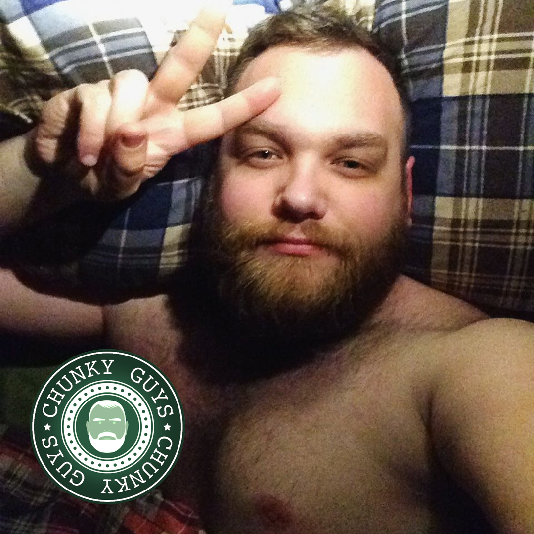 Younger bearded man lying in bed, one hand showing a peace sign, looking up at the camera