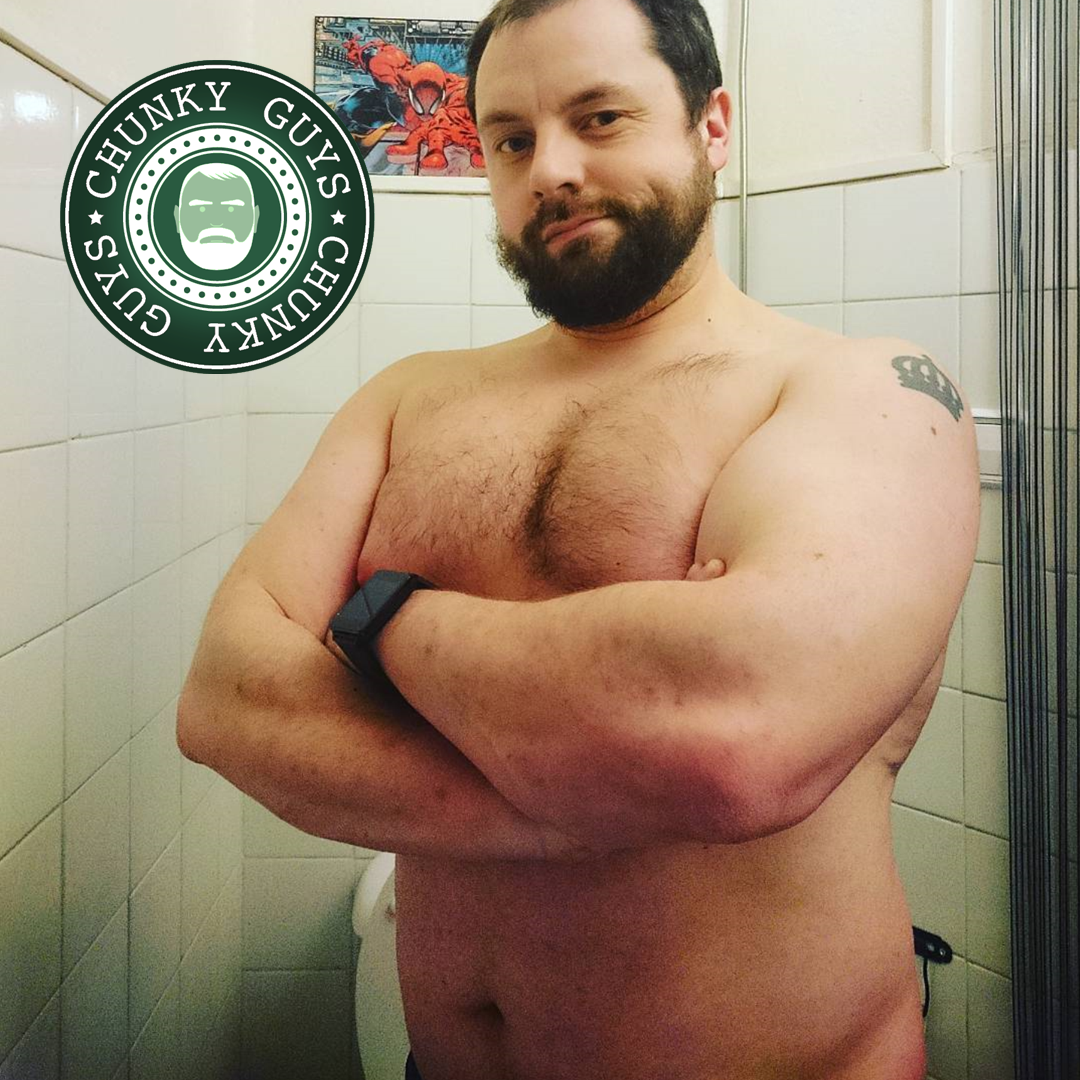 Bearded chunky guy with hairy pecs standing with his arms folded looking at the camera