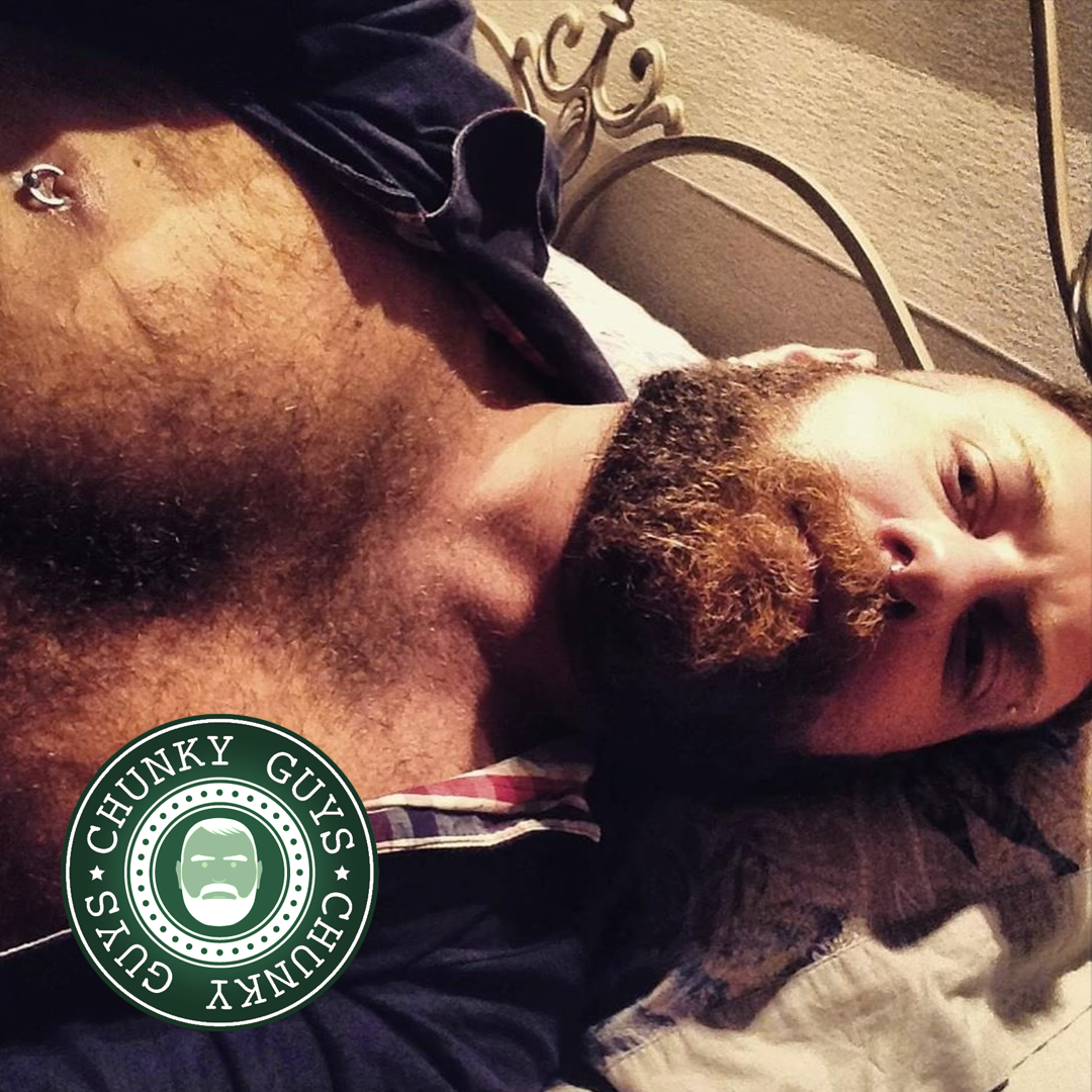 Bearded guy with a hairy chest and nipple piercing lying down looking up at the camera