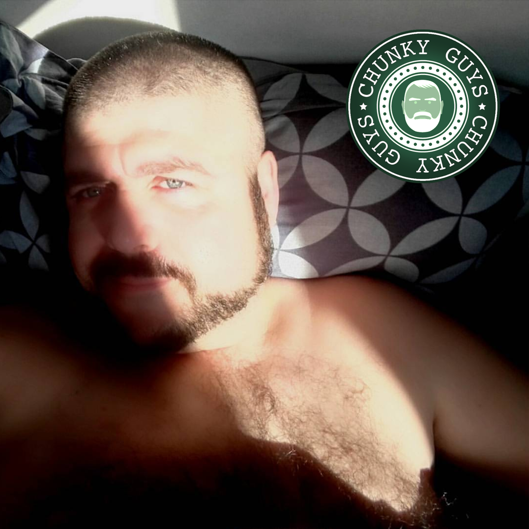 Beary bearded guy with short hair and a hairy chest lying in bed with the sun shining across his face