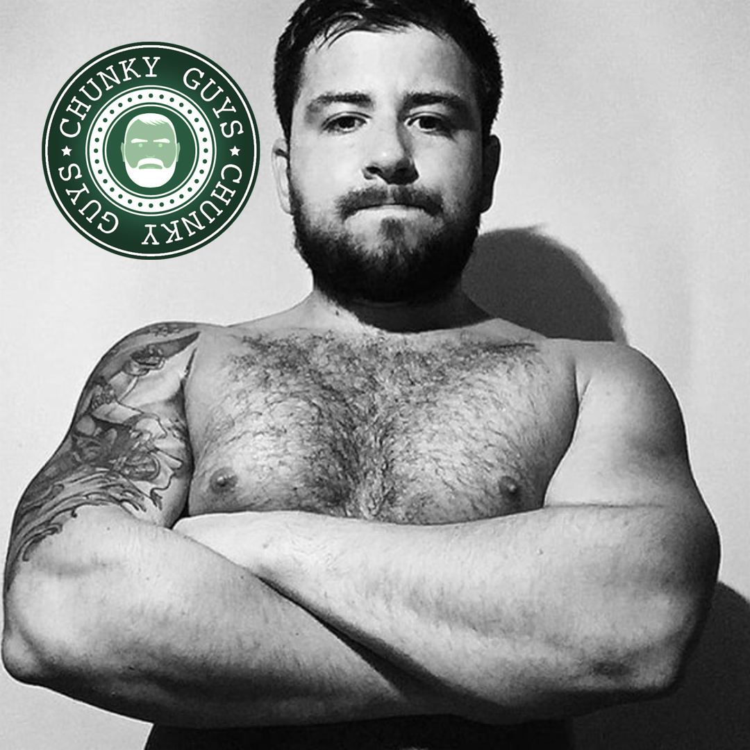 Young tattooed guy with a hairy chest and a beard stands with his arms folded