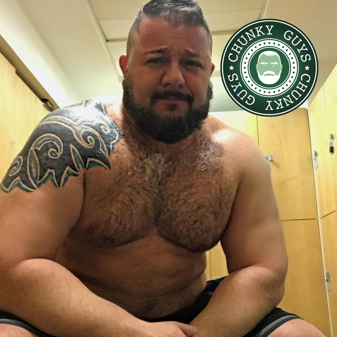 Tattooed and tanned muscle bear with dark hair and a beard wearing shorts sitting in a changing room