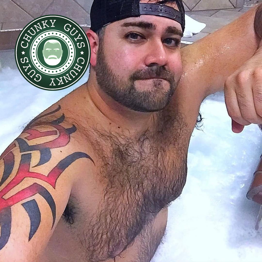 Tattooed hairy guy wearing a baseball cap looking up at the camera and standing in a hot tub