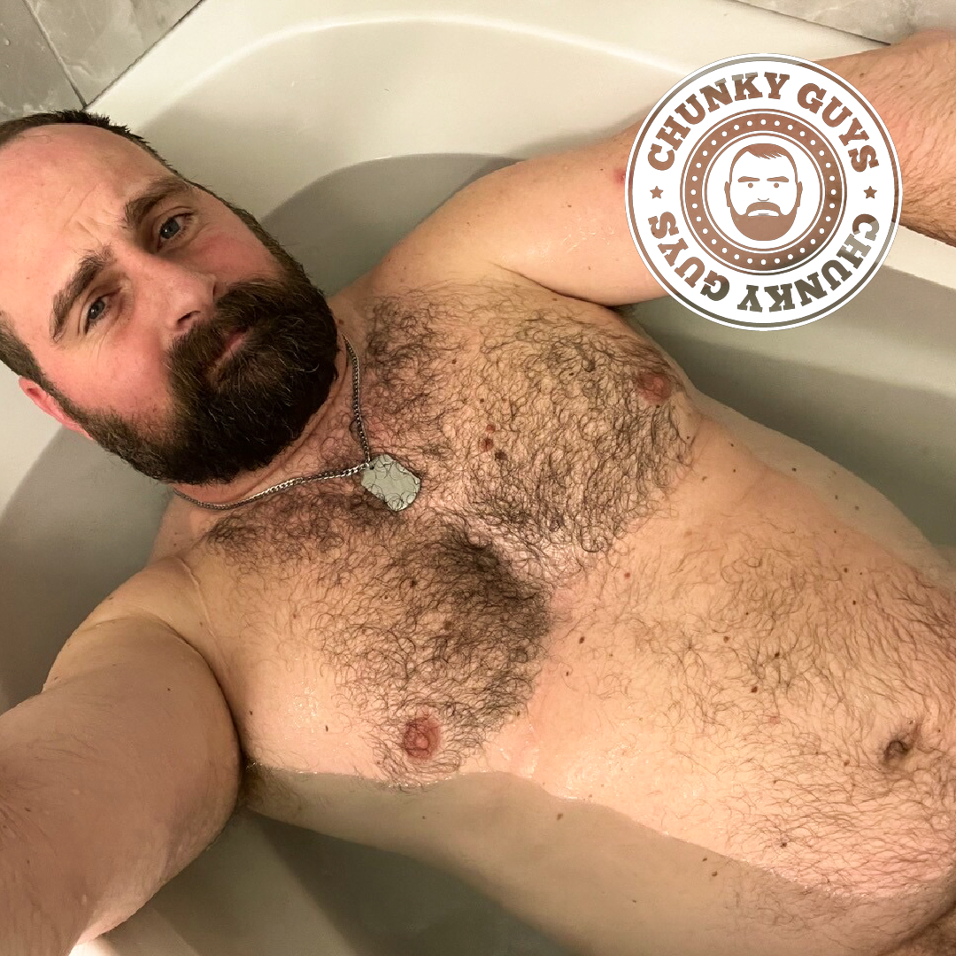 Chunky muscled bearded bear with a hairy chest lying in a bath wearing dog tags around his neck