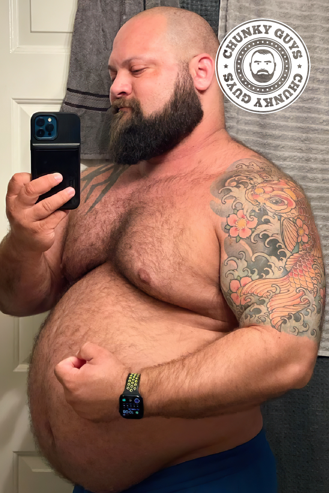 Chunky muscle bear with a round belly and tattoos poses in front of the mirror to take a photo
